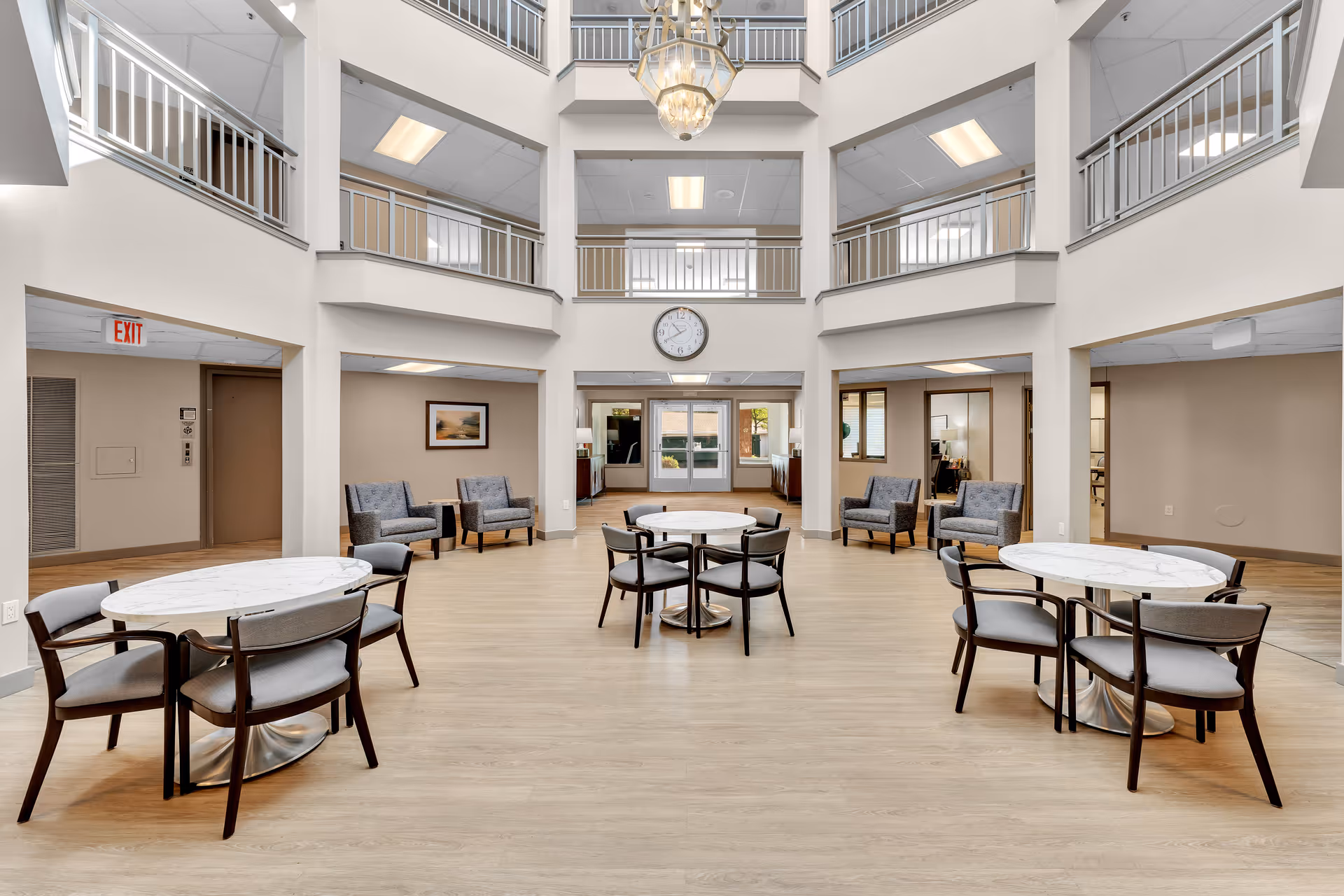 Two-story atrium-style common area with round dining tables and chairs, lounge seating, a central clock and chandelier.