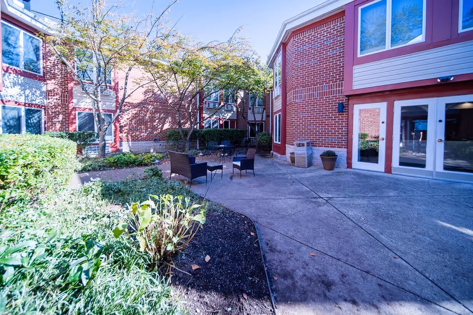 Outdoor courtyard area at Independence Court of Hyattsville featuring brick buildings with multiple windows, a concrete patio with outdoor chairs and tables, surrounded by greenery and trees under a clear sky.