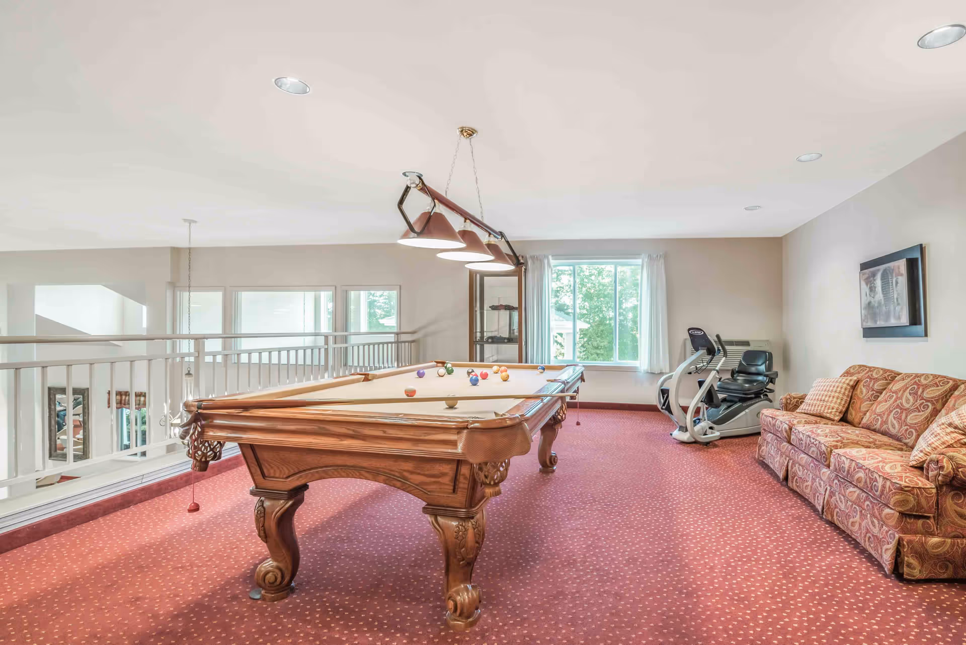 A carpeted recreation room with a wooden pool table, overhead lights, a patterned sofa and exercise equipment by a window.