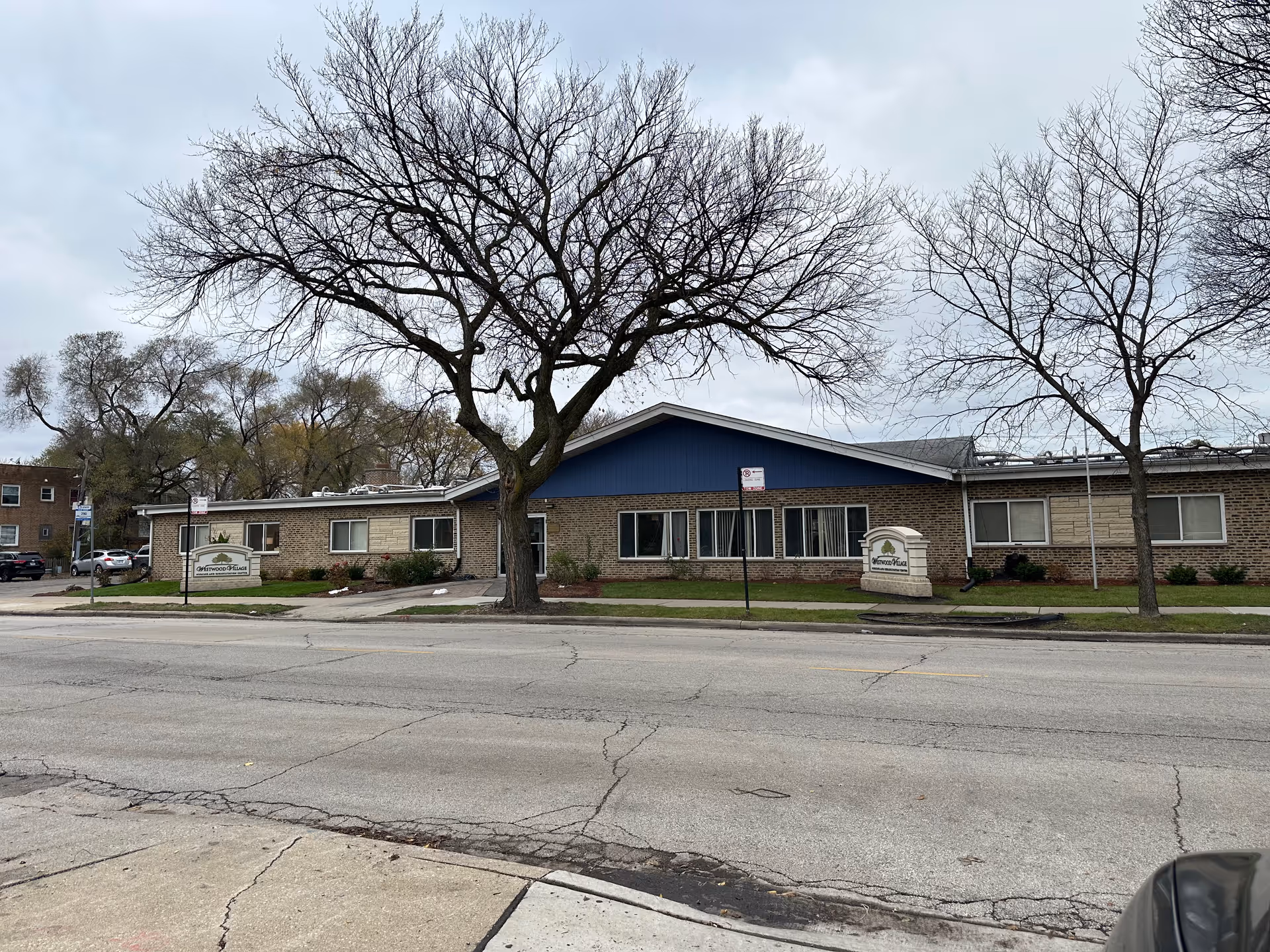 Exterior view of Westwood Village Nursing and Rehabilitation Center, a single-story brick building with a blue gable roof section. There are leafless trees in front of the building and two signs displaying the facility's name. The street and sidewalk are visible in the foreground under a cloudy sky.