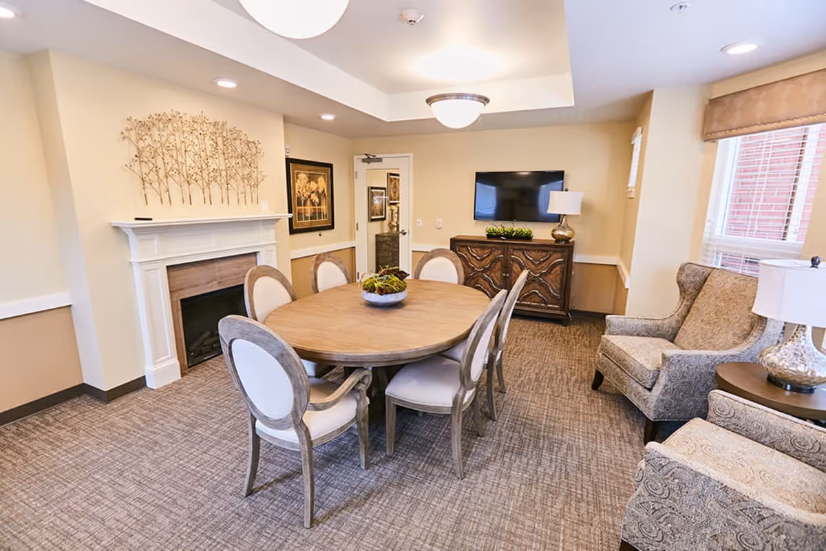 A cozy interior room with a round wooden table surrounded by six upholstered chairs. The room features a white fireplace with decorative metal wall art above it, a wooden cabinet with a flat-screen TV mounted on the wall above, and two patterned armchairs near a window with blinds. The room is warmly lit with ceiling lights and a table lamp.
