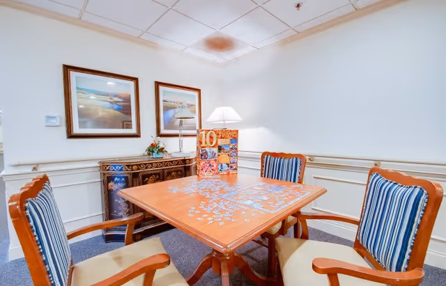 A small room with a wooden table in the center, four wooden chairs with blue and white striped cushions around it. A partially completed jigsaw puzzle is spread out on the table. Against the wall is a decorative cabinet with a lamp and a photo collage on top. Two framed landscape paintings hang on the wall above the cabinet. The room has white walls and a drop ceiling with fluorescent lighting.