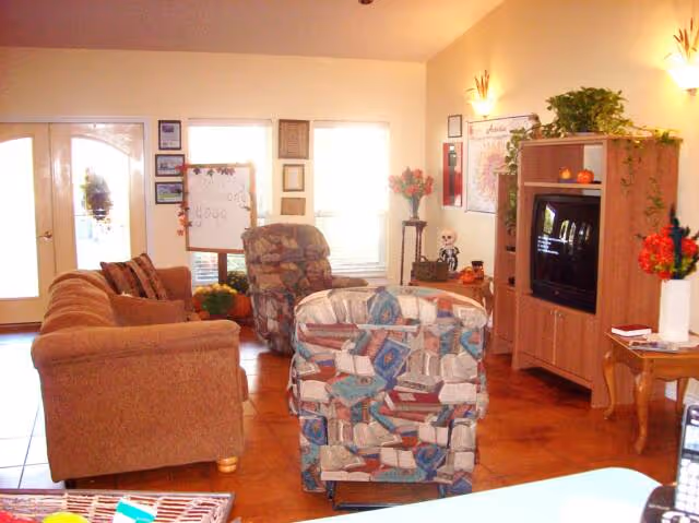 A cozy living room with a brown sofa, a patterned armchair, and a recliner facing a wooden entertainment center with a television. The room has tile flooring, wall-mounted lights, plants on top of the entertainment center, and various decorations including framed pictures and flowers. There is a door and two windows letting in natural light.