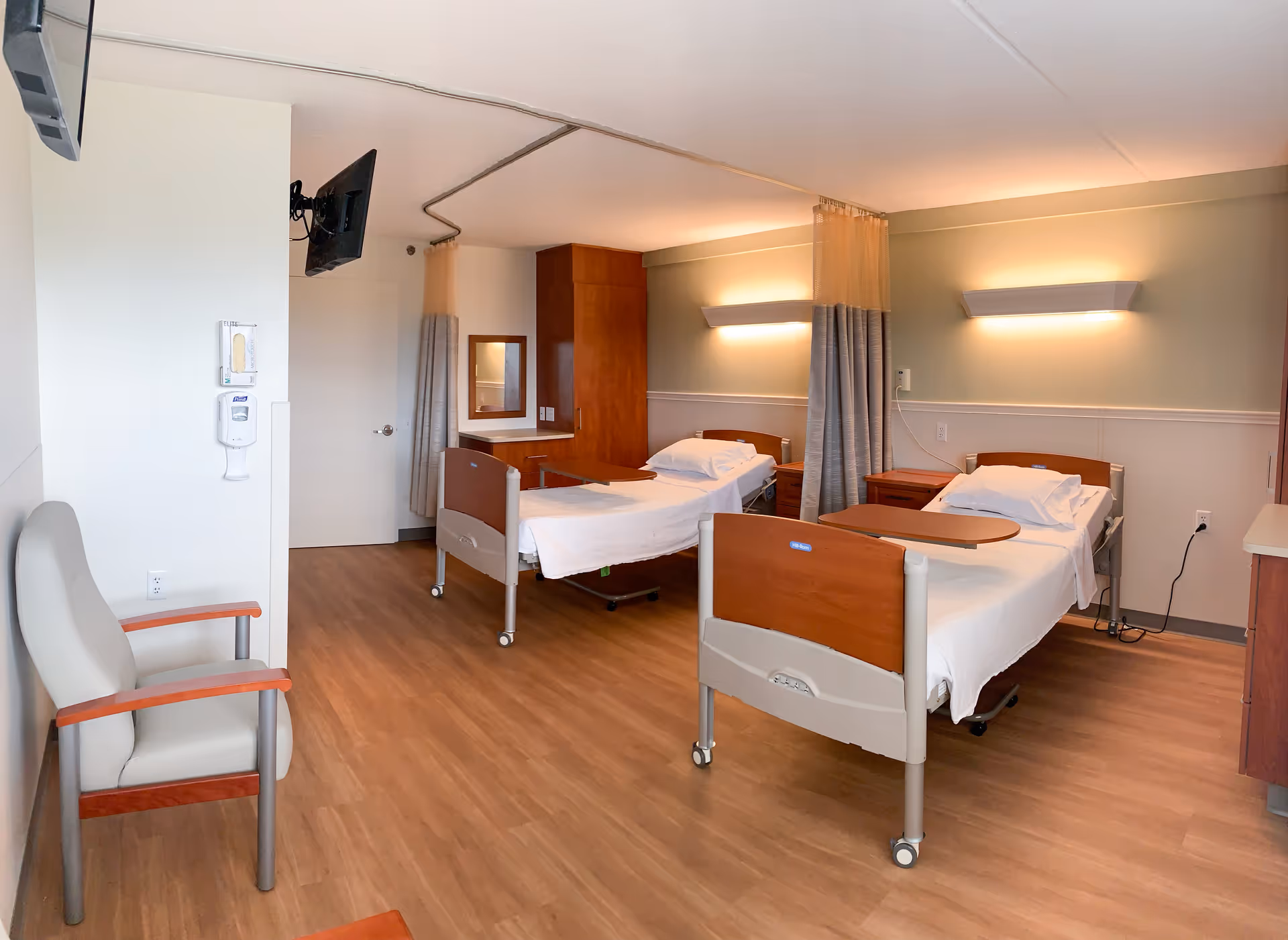 A clean and well-lit nursing facility room with two hospital beds, each equipped with a white pillow and sheets. The beds have wooden headboards and footboards with attached overbed tables. There are privacy curtains between the beds and wall-mounted lights above each bed. A beige armchair with wooden armrests is positioned near the left wall, which also has a mounted hand sanitizer dispenser and a television. The floor is covered with wood-patterned vinyl flooring, and there is a wooden cabinet and mirror in the back corner of the room.
