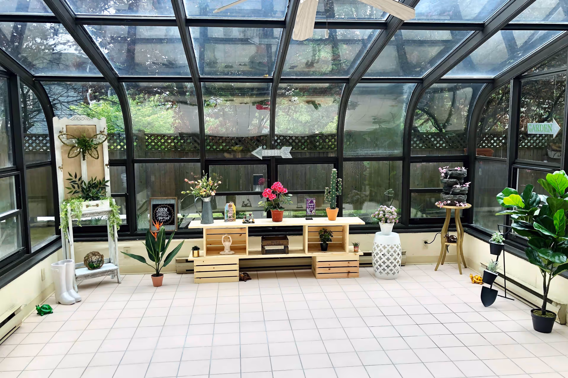 Bright glass-enclosed sunroom with tiled floor, potted plants and wooden shelving displaying flowers and decor.