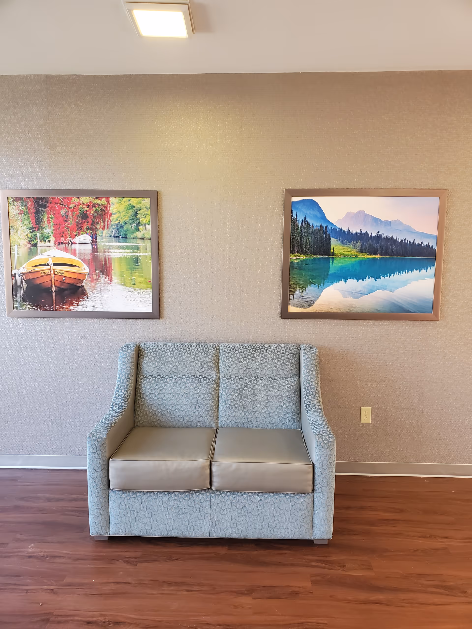 A small upholstered two-seater sofa against a textured wall with two framed landscape pictures above it.