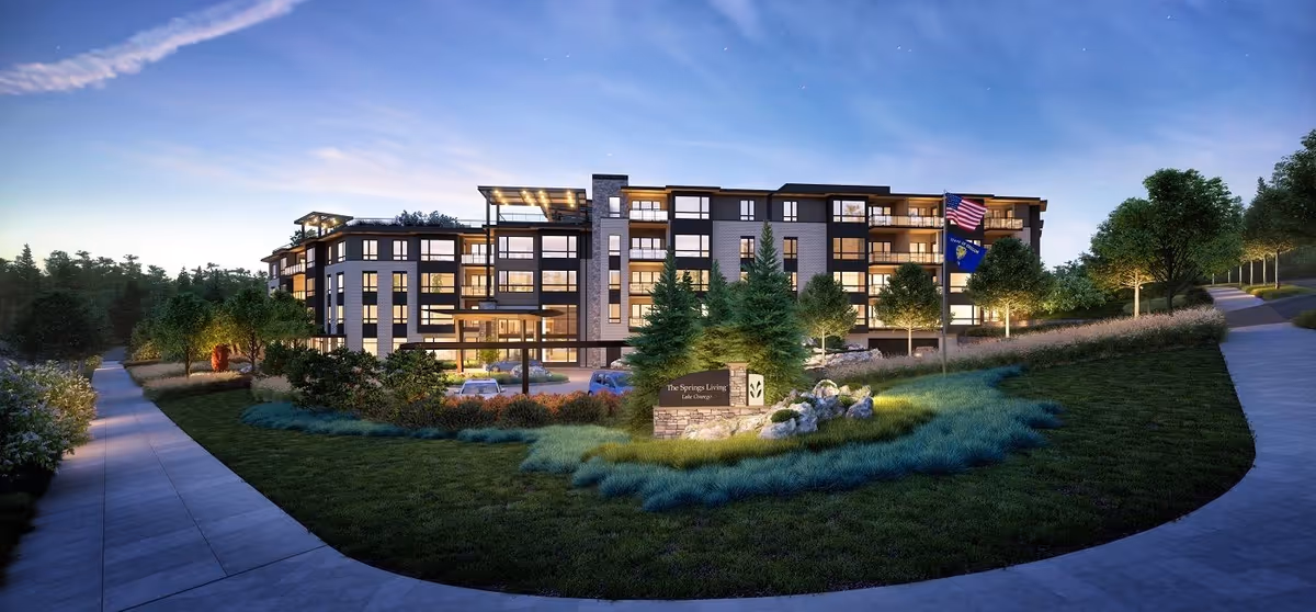 Exterior view of a multi-story senior living building at dusk with landscaped grounds, a circular driveway and illuminated windows.