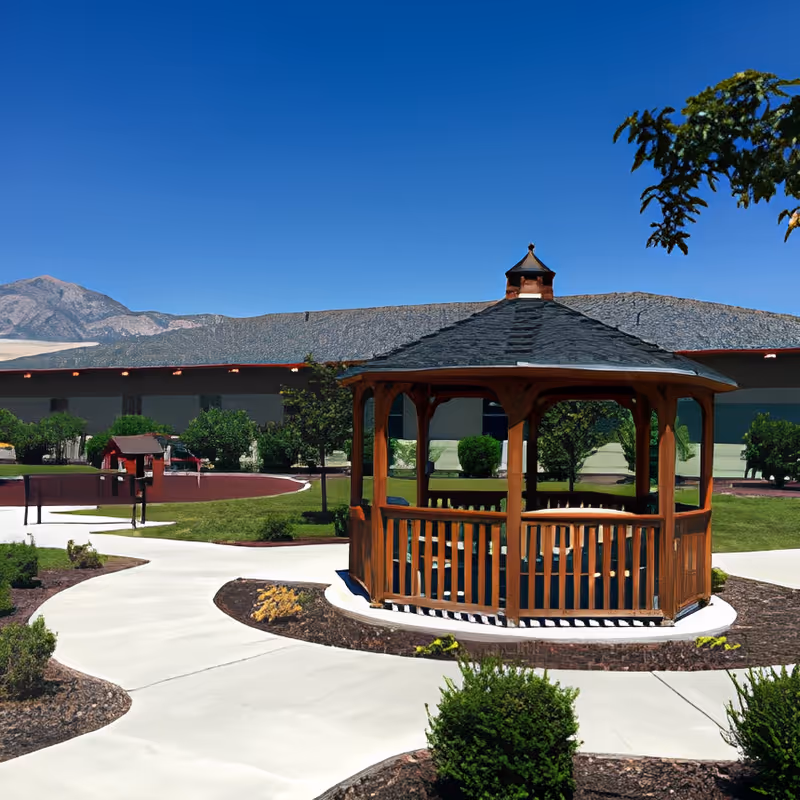 Outdoor scene at George E. Wahlen Ogden Veterans Home featuring a wooden gazebo with a shingled roof surrounded by paved walkways, green grass, small bushes, and trees under a clear blue sky with mountains in the background.