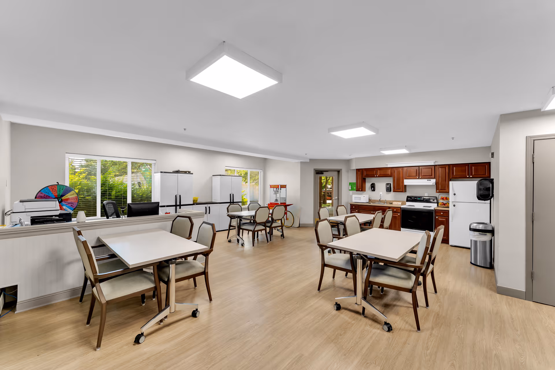 Bright communal dining and activity room with several tables and chairs, a kitchenette and refrigerators along the back wall.
