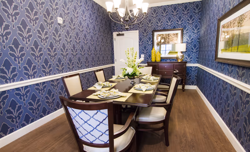 A dining room with a dark wooden table set for six people. The chairs have cream-colored cushions and patterned backs. The walls are covered with blue wallpaper featuring a decorative pattern, and there is white chair rail molding. A chandelier hangs above the table, and a sideboard with yellow vases and a lamp is against the far wall. Two framed pictures hang on the walls.