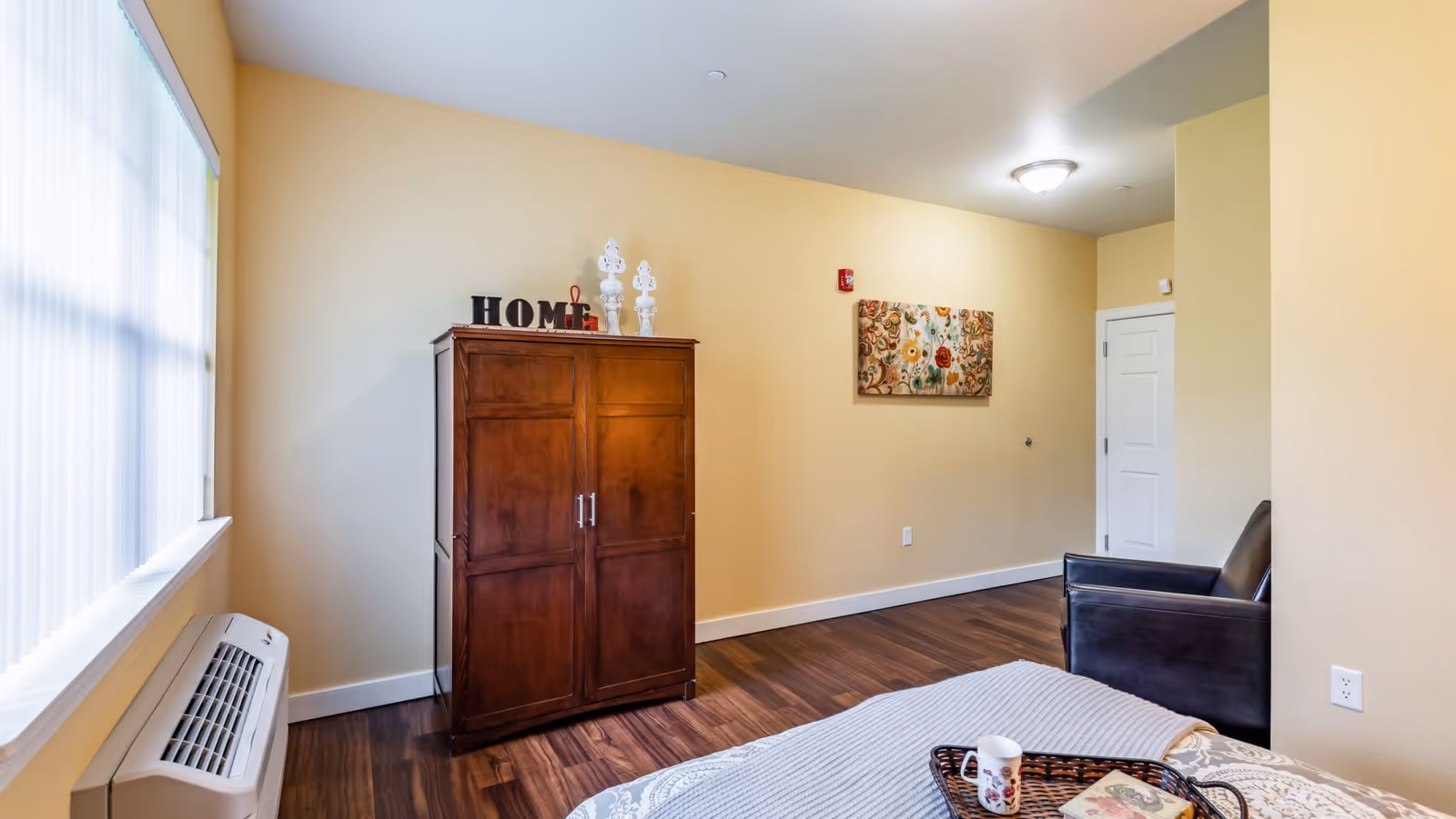 A bedroom with light yellow walls and wooden flooring. There is a wooden cabinet with decorative items and the word 'HOME' on top, a black armchair, and a bed with a tray holding a cup and a book. A window with blinds and an air conditioning unit is on the left side, and a floral painting hangs on the wall.