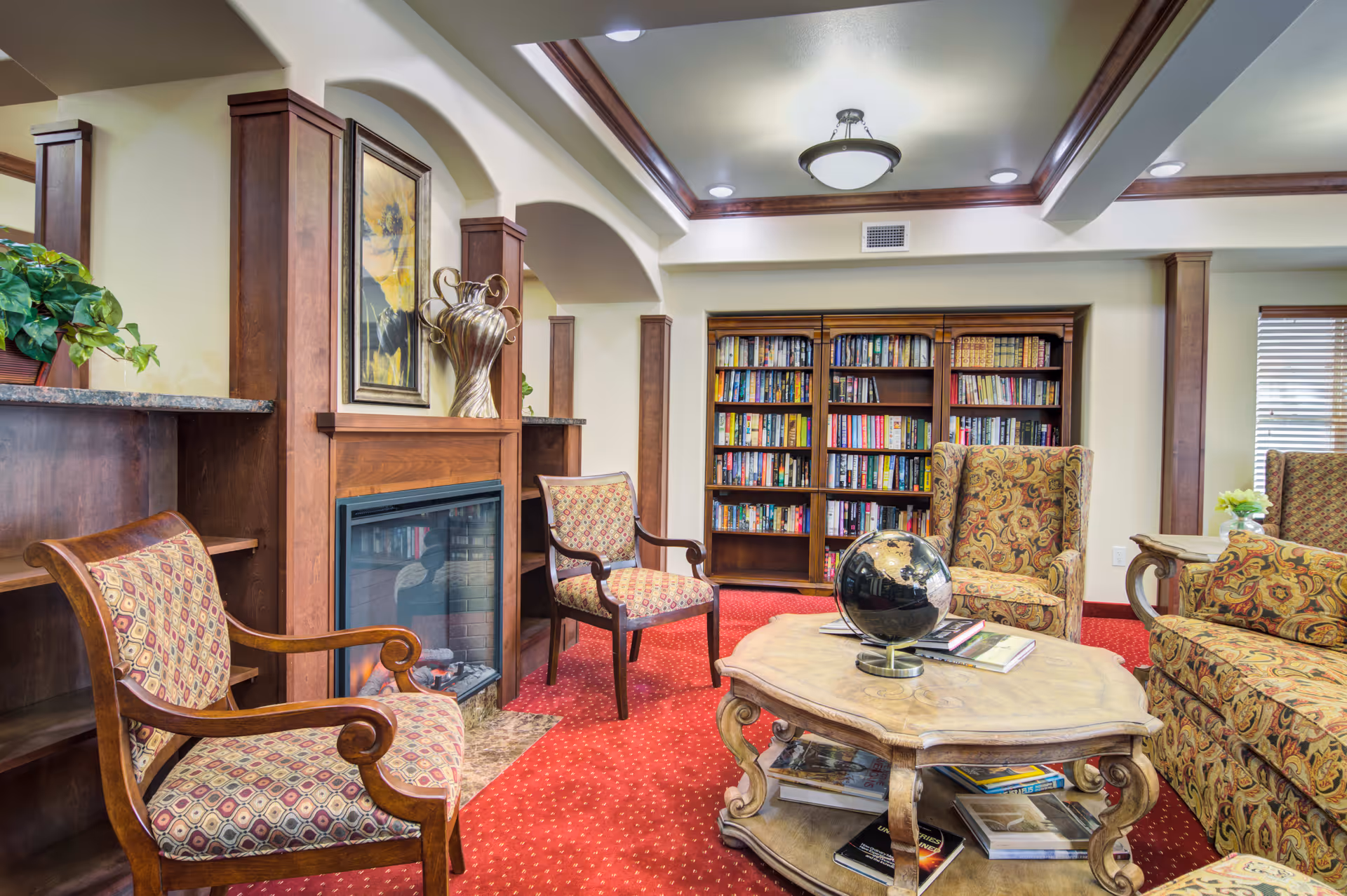 A cozy living room area with patterned upholstered chairs and a sofa arranged around a wooden coffee table with books and a globe on it. The room features a fireplace with a decorative vase and framed artwork above it, built-in wooden bookshelves filled with books, and a red carpeted floor. The ceiling has recessed lighting and a central light fixture.