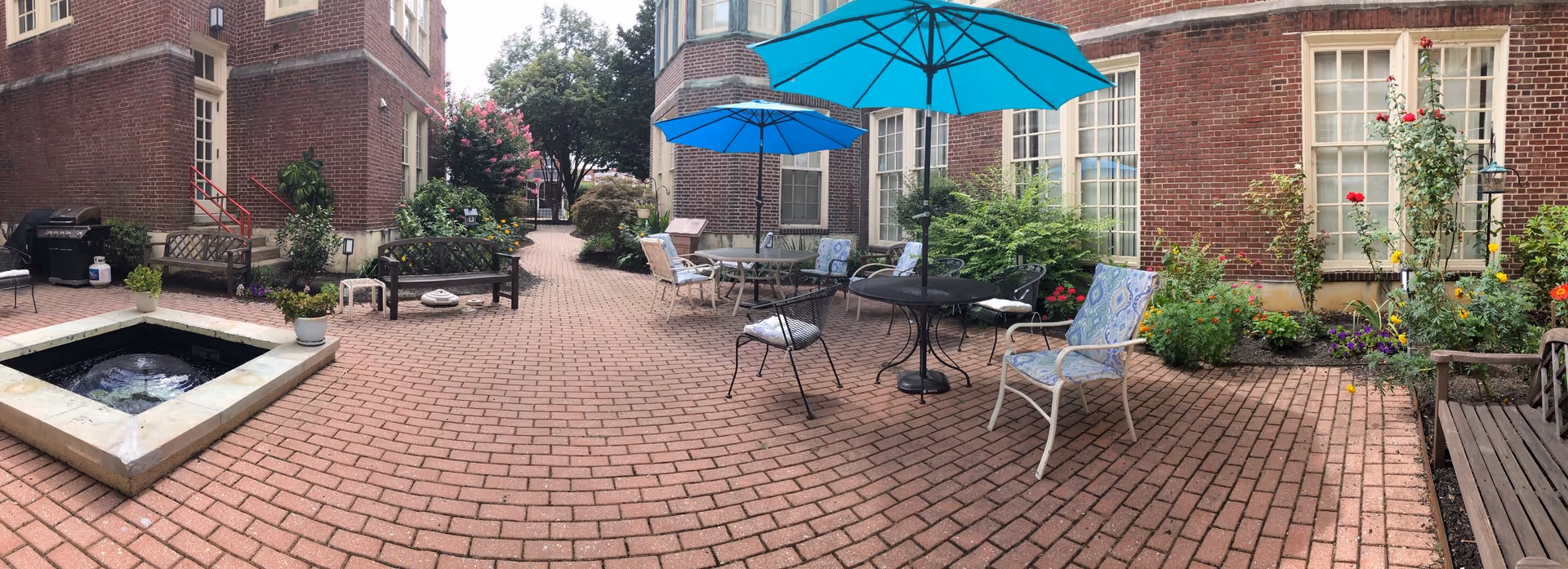 A brick-paved outdoor courtyard area between two brick buildings, featuring several patio tables with blue umbrellas, chairs with cushions, benches, potted plants, and a small square water fountain. There are various green shrubs and flowering plants along the edges of the courtyard.