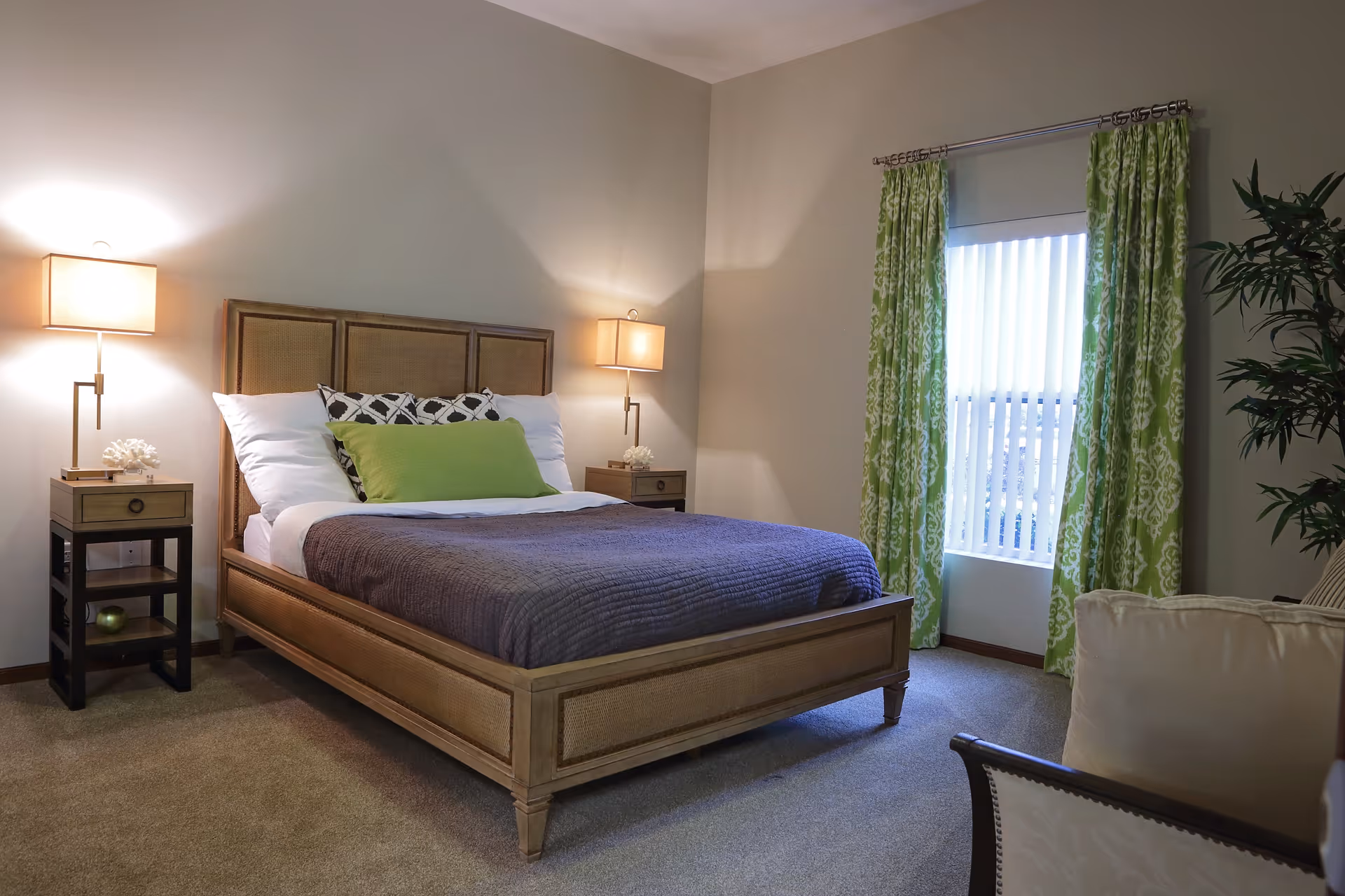 Bedroom with a wooden bed dressed with pillows and a gray blanket, flanked by nightstands and lamps, green patterned curtains, and a chair.