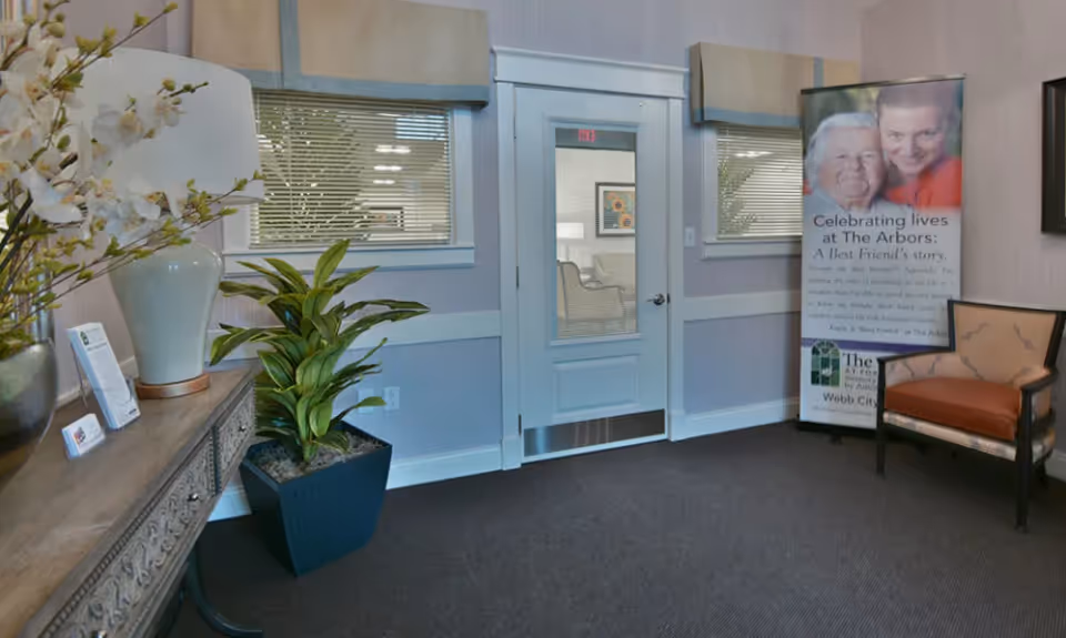 Interior view of a senior living facility hallway with a closed white door featuring a glass panel, two windows with blinds on either side, a decorative wooden console table with a white lamp and flower arrangement, a potted plant, a cushioned chair, and a standing banner promoting The Arbors at Foxberry Terrace.