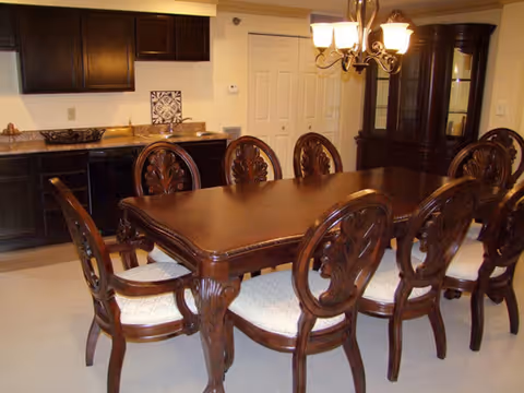 A dining room with a large wooden dining table surrounded by eight ornate wooden chairs with cushioned seats. In the background, there are dark wooden cabinets, a countertop, a chandelier with five lights hanging above the table, and a glass-fronted china cabinet.