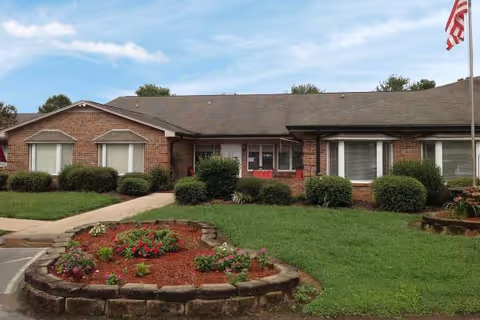 A single-story brick senior living facility with manicured lawn and circular flower bed.