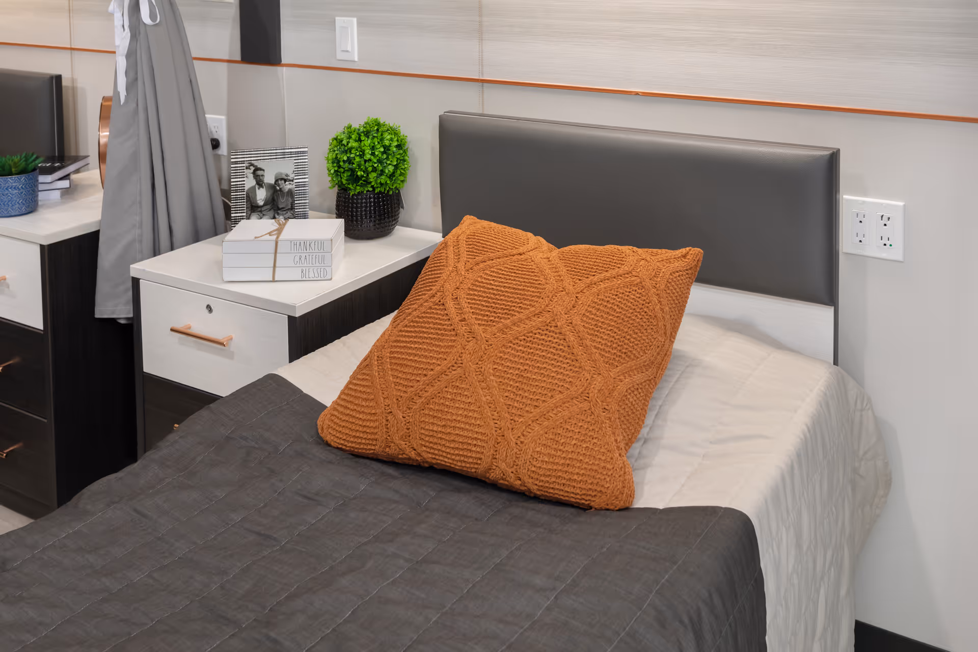 A neatly made single bed with a gray headboard, white and gray bedding, and a large textured orange pillow. Next to the bed is a nightstand with a small green plant, a framed black and white photo, and a decorative block with the words 'Thankful Grateful Blessed'. A gray curtain and part of a dresser are visible in the background.