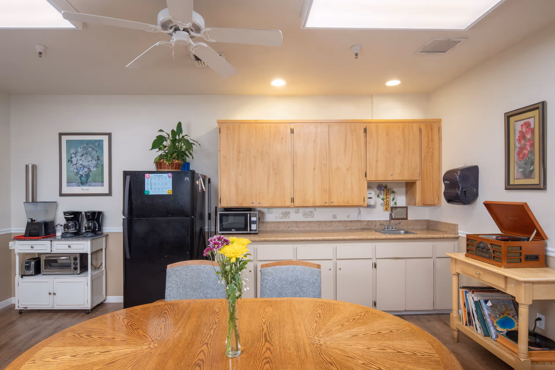 A small kitchen area in an assisted living facility featuring wooden cabinets, a black refrigerator, a microwave, a coffee maker, and a toaster oven. A round wooden table with a vase of colorful flowers is in the foreground. The room has framed floral artwork on the walls, a ceiling fan, and a wooden side table with a record player and books underneath.