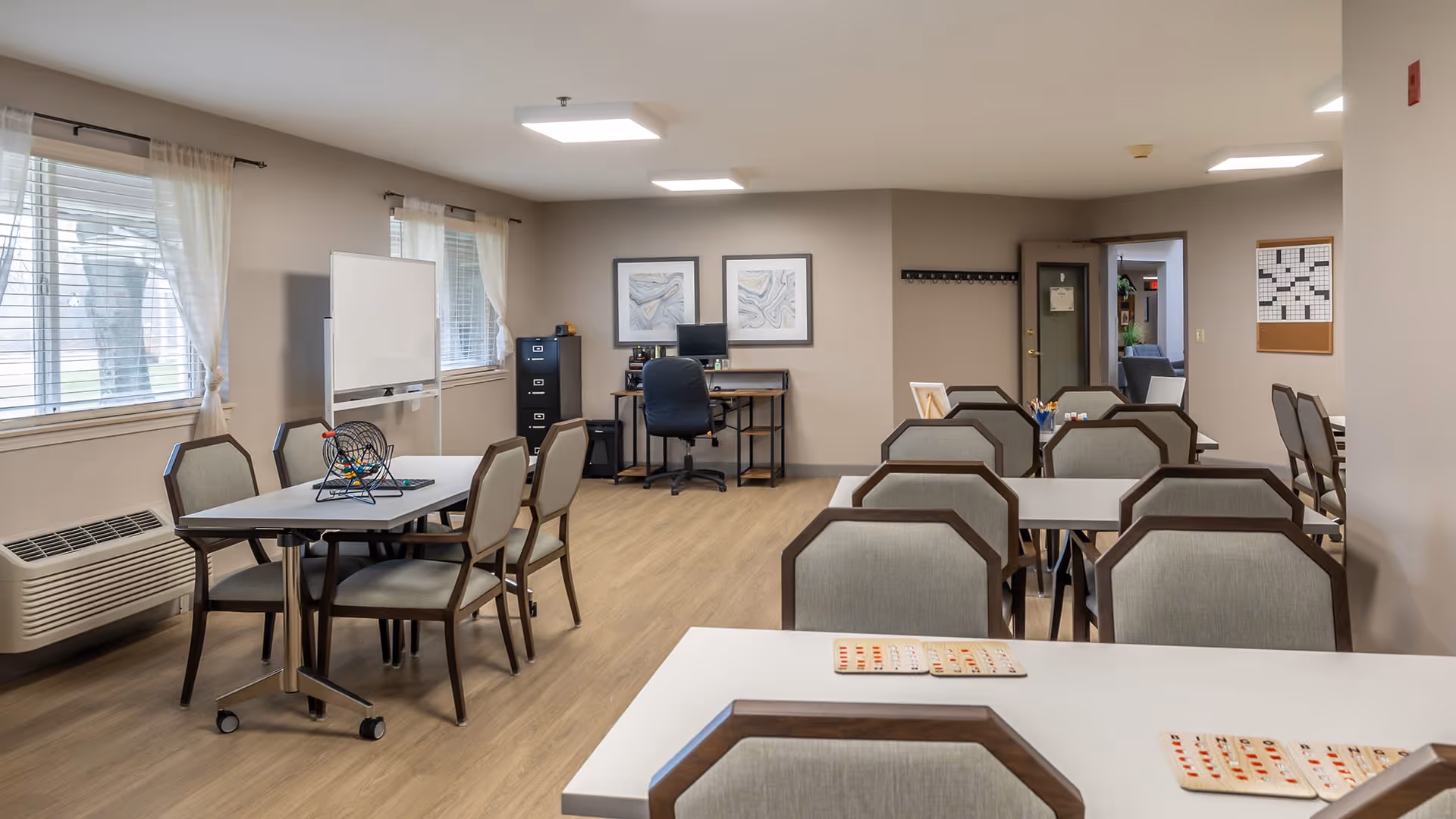 A well-lit common room with multiple tables and chairs arranged for group activities. There are bingo cards and a bingo cage on the tables, a whiteboard near the windows with sheer curtains, a desk with a computer and filing cabinet against the far wall, and a crossword puzzle board on the wall near a doorway.