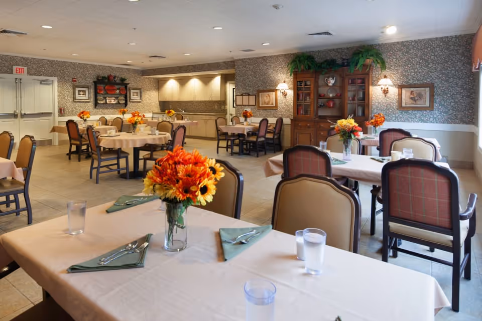 A dining room in Cedar Crest Alzheimer's Special Care Center with several tables covered in beige tablecloths, each set with green napkins, silverware, glasses of water, and floral centerpieces featuring orange and yellow flowers. The room has floral wallpaper, a wooden cabinet with decorative items, and soft lighting from wall sconces. There are multiple chairs around each table and a kitchen area visible in the background.