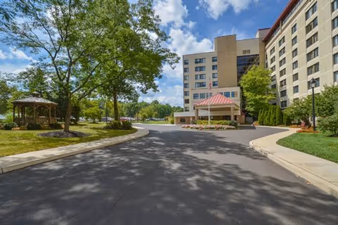 Exterior view of a multi-story senior living facility with a driveway leading to the main entrance. There is a gazebo on the left side surrounded by trees and greenery under a partly cloudy sky.