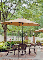 Outdoor patio area with metal chairs and tables under large beige umbrellas surrounded by trees and greenery, with a white fence and brick pillars in the background.