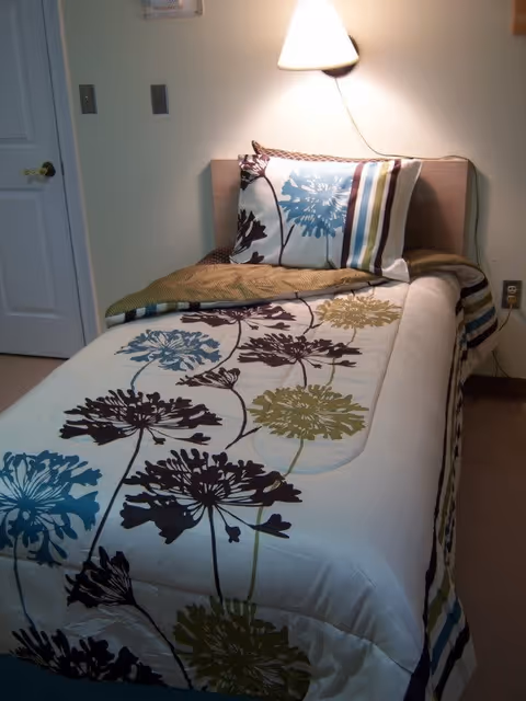 A single bed with a floral patterned comforter and matching pillow in a small room. A wall-mounted lamp above the headboard illuminates the bed. There is a closed white door to the left and an electrical outlet on the wall to the right.