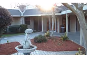 Outdoor courtyard area of a nursing and rehabilitation facility with a small statue fountain in the center, surrounded by mulch and plants, with a covered walkway and building in the background.