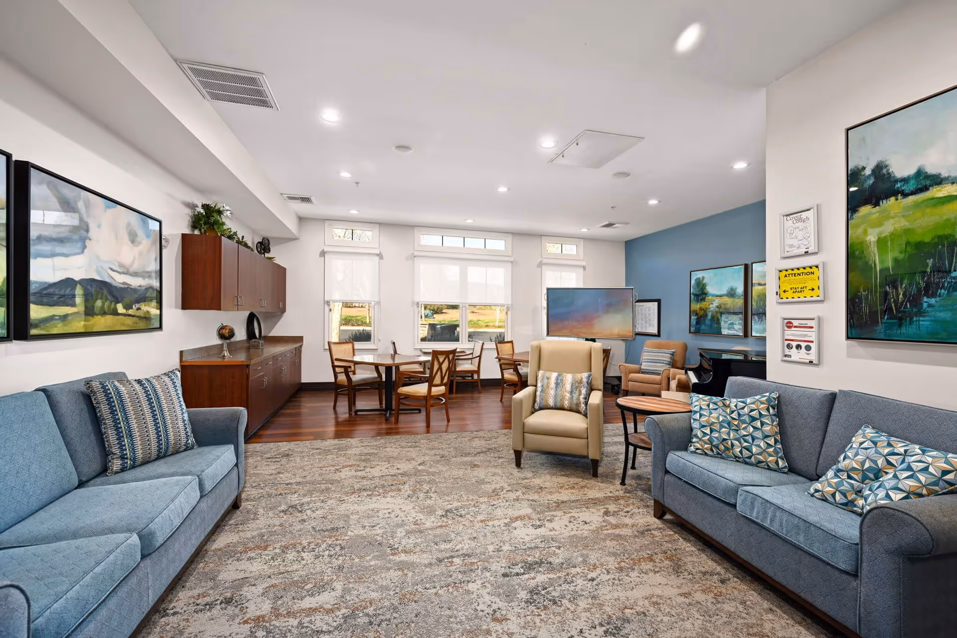 A bright and spacious living room area in Sunol Creek Memory Care featuring two blue sofas with patterned pillows, a beige armchair with a decorative pillow, a round wooden side table, and a large area rug. The room has large windows with white blinds, wooden flooring, and several landscape paintings on the walls. There is a wooden cabinet along one wall and a TV on a stand near the back.