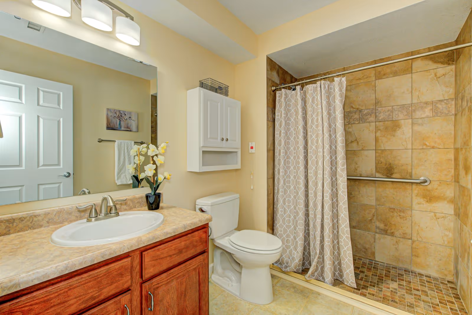 A clean and well-lit bathroom featuring a sink with a marble countertop and wooden cabinet, a toilet, and a tiled walk-in shower with a patterned shower curtain and a grab bar. Above the toilet is a white wall-mounted cabinet, and a large mirror with three overhead lights is visible above the sink. A small vase with yellow flowers is placed on the countertop.