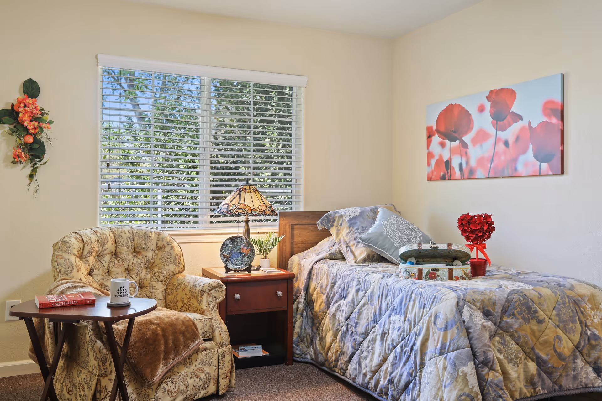A cozy senior living bedroom with a single bed covered in a patterned quilt and decorative pillows. Next to the bed is a wooden nightstand with a decorative lamp, a small plant, and a round decorative plate. A comfortable upholstered armchair with a floral pattern sits beside a small wooden table holding a book and a coffee mug. A window with white blinds lets in natural light, and a wall decoration with red flowers hangs above the bed.