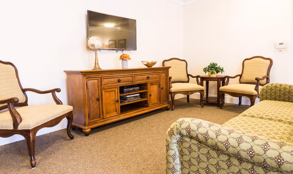 A cozy living room area with a patterned green sofa in the foreground, three wooden armchairs with beige cushions, a wooden TV stand with decorative items including a globe and a vase with orange flowers, and a flat-screen TV mounted on a white wall.