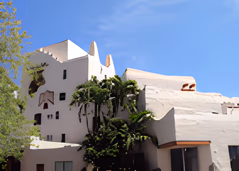 Exterior view of a uniquely designed building with white stucco walls, irregular shapes, and small windows, surrounded by trees and set against a clear blue sky.