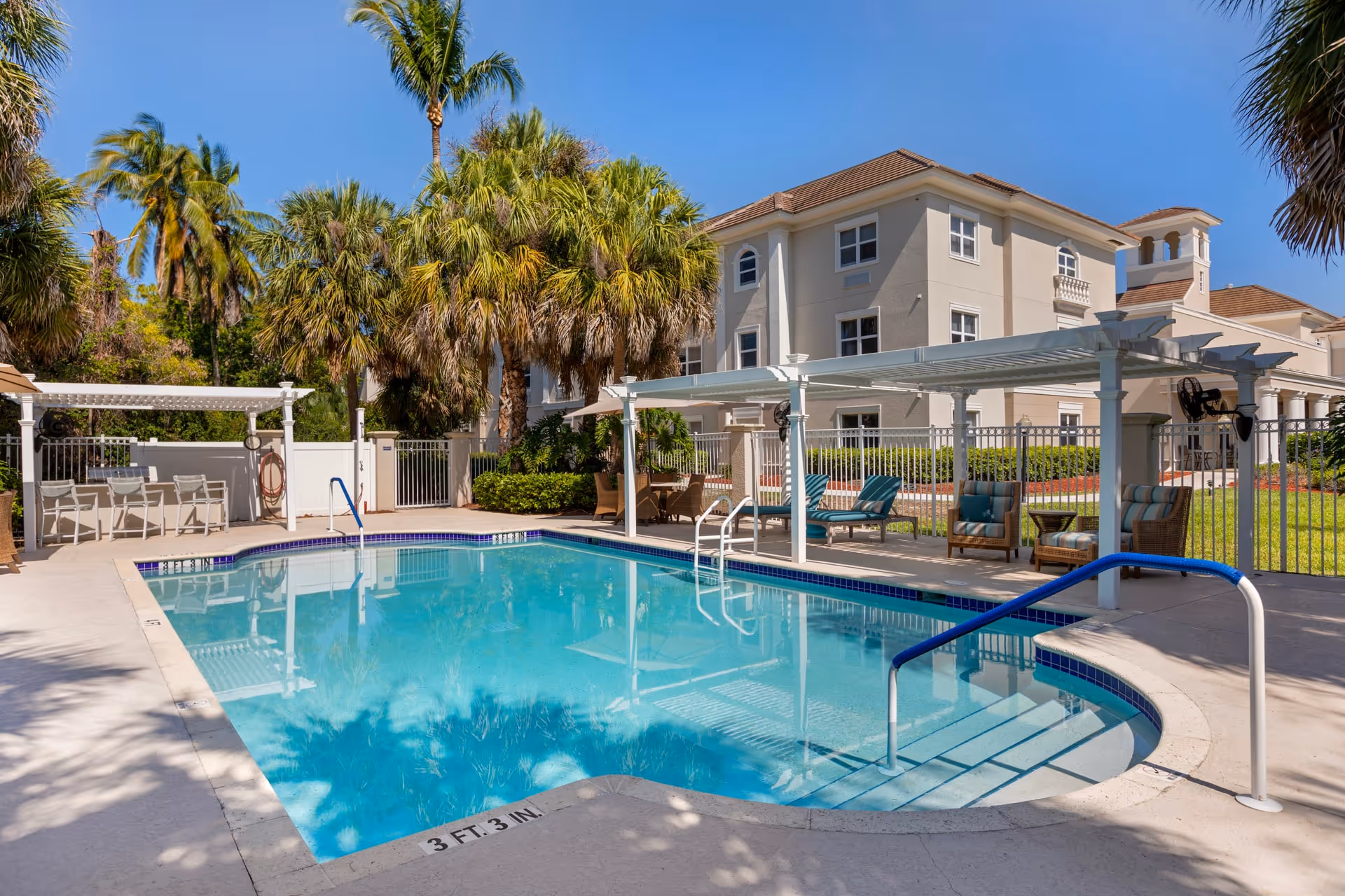 Outdoor swimming pool area at Brookdale Bonita Springs with clear blue water, surrounded by palm trees and lounge chairs under white pergolas. A beige multi-story building is visible in the background under a clear blue sky.