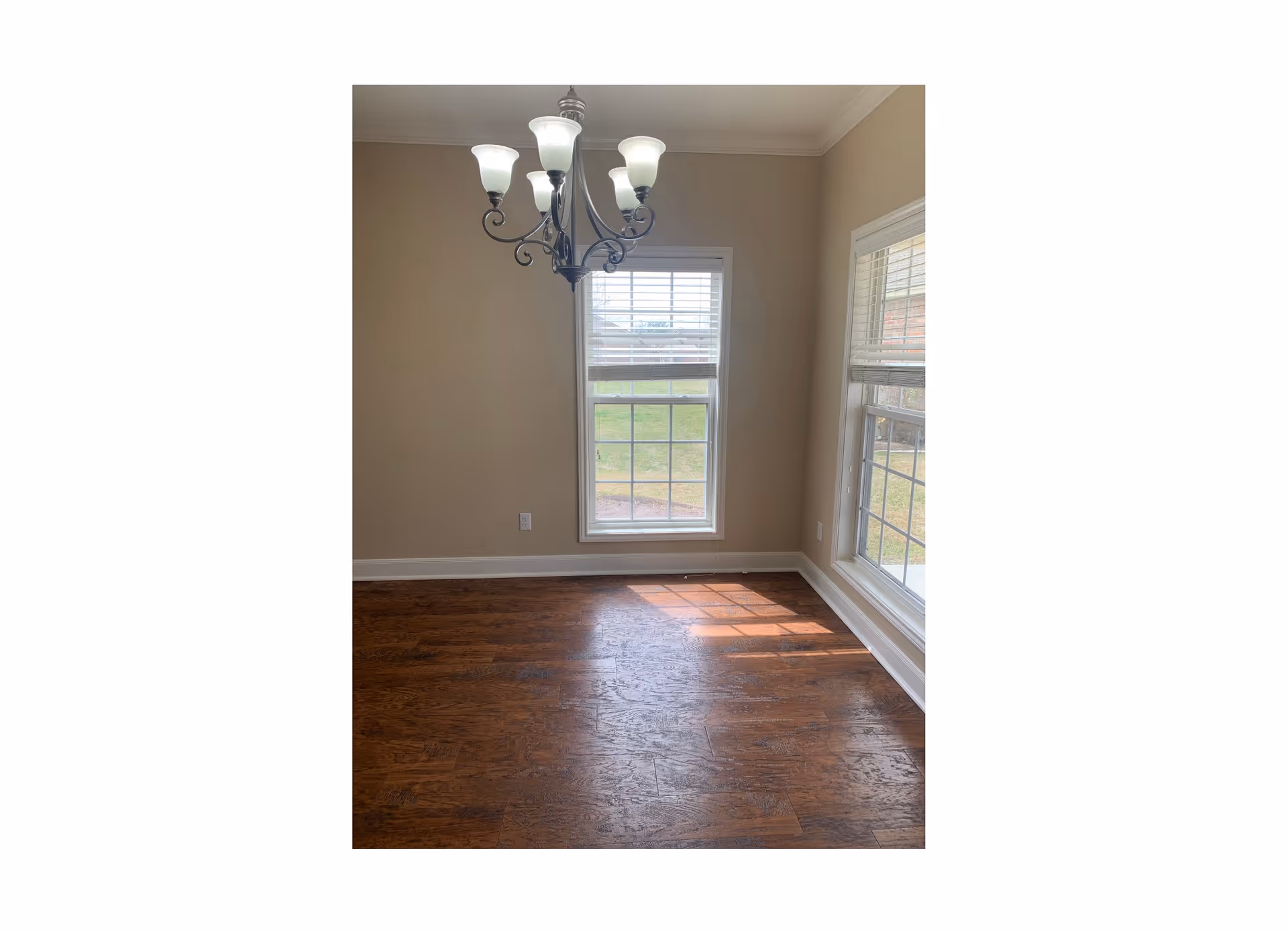 Empty dining area with hardwood floors, a decorative chandelier, and two tall windows letting in sunlight.