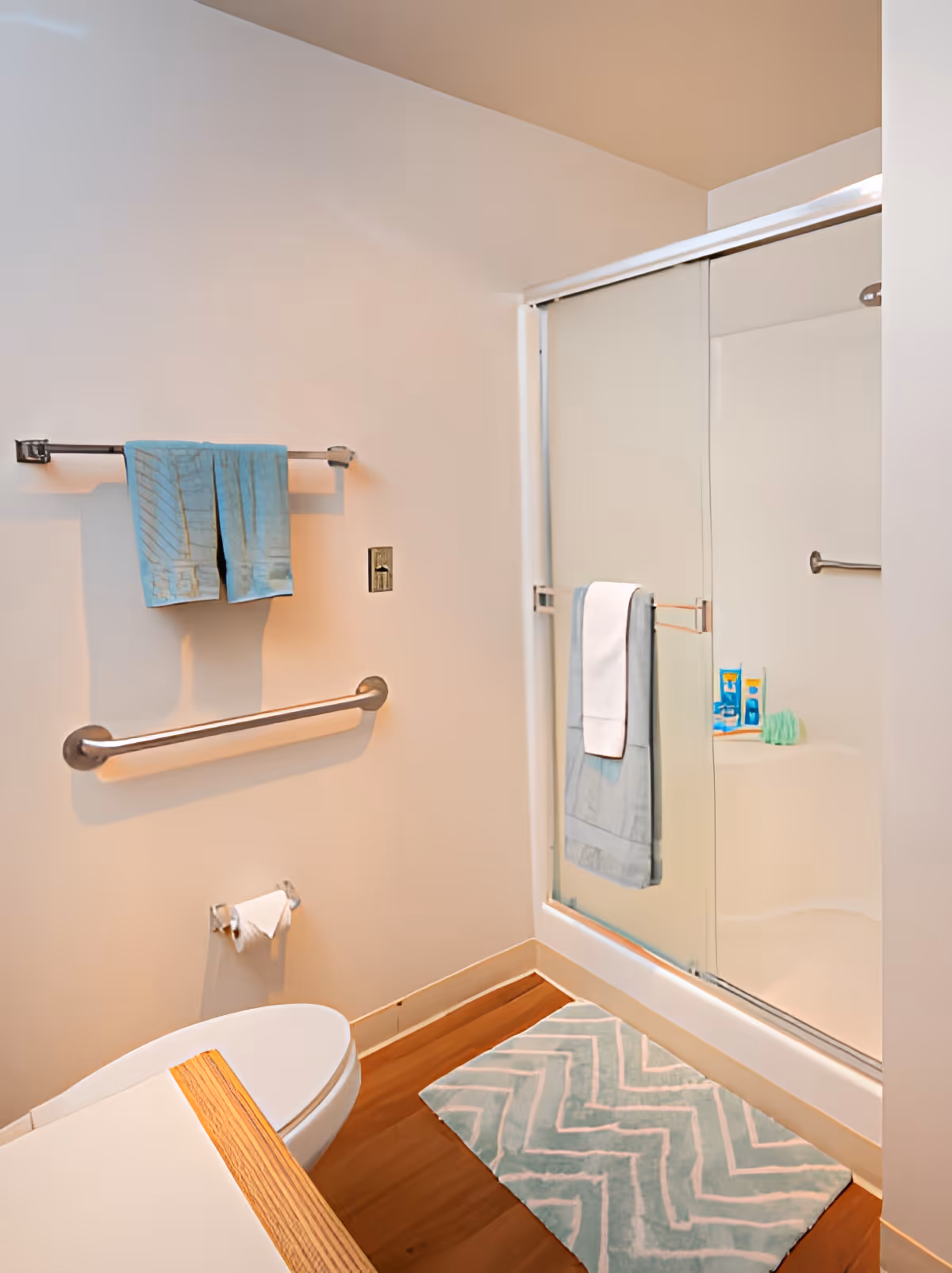 A clean bathroom with a white toilet, a wooden countertop, a towel rack with blue towels, a grab bar on the wall, a toilet paper holder, a shower with sliding glass doors, and a blue and white chevron-patterned bath mat on the wooden floor.