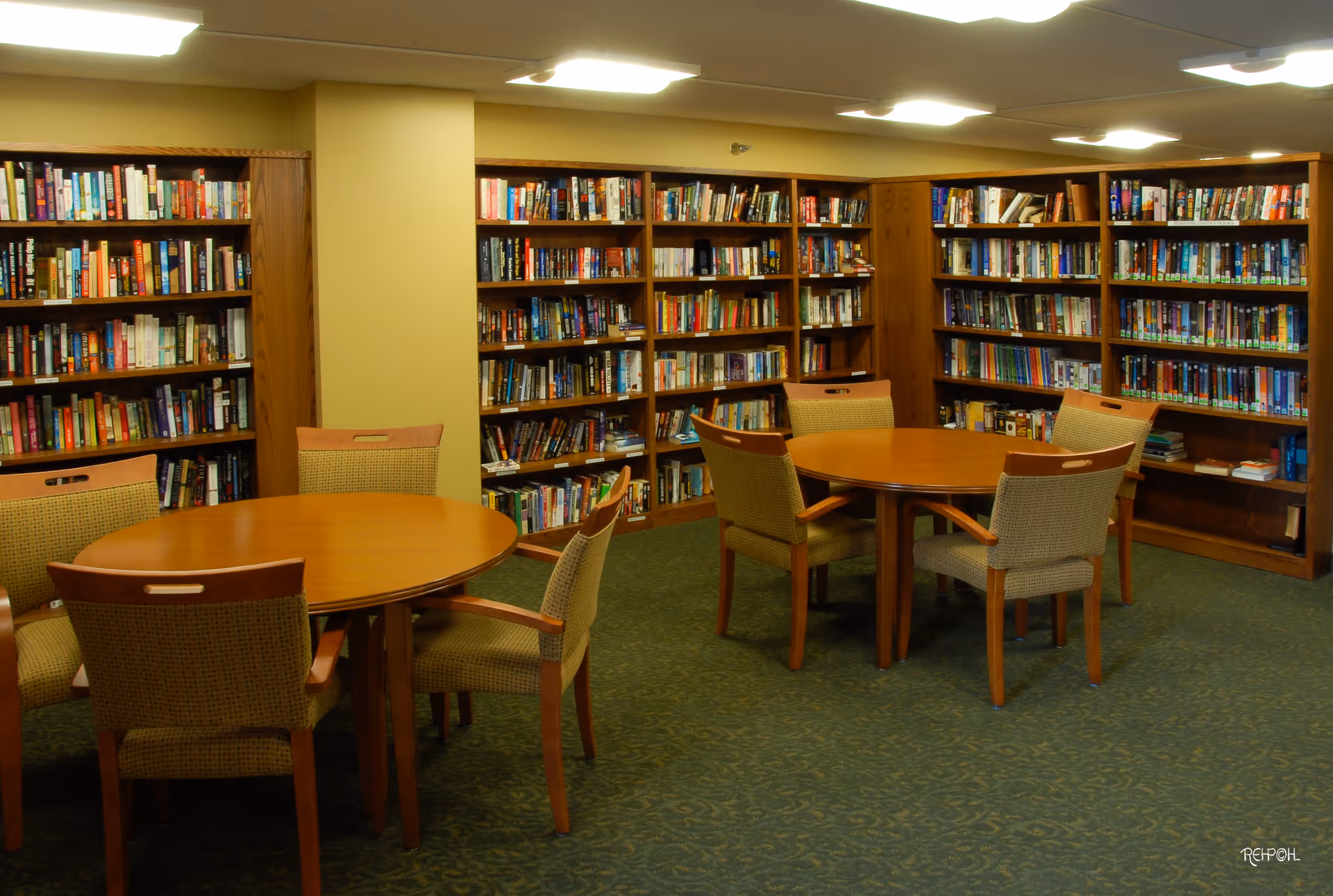 A cozy library room with wooden bookshelves filled with books lining the walls. There are two round wooden tables, each surrounded by four cushioned chairs with wooden frames. The room has a green patterned carpet and soft overhead lighting.