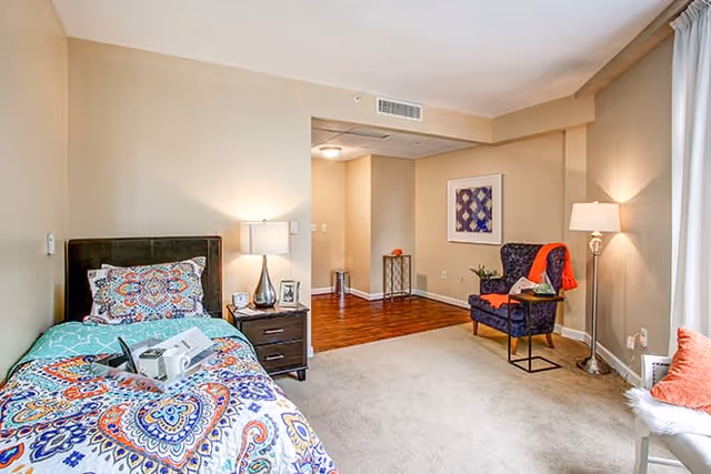 A bright and cozy bedroom in a senior living facility with a single bed covered in a colorful patterned bedspread, a nightstand with a lamp and framed photos, a comfortable armchair with an orange throw blanket, a floor lamp, and a window with sheer curtains letting in natural light. The room has beige walls and carpeted flooring, with a small open area leading to a hardwood floor section.