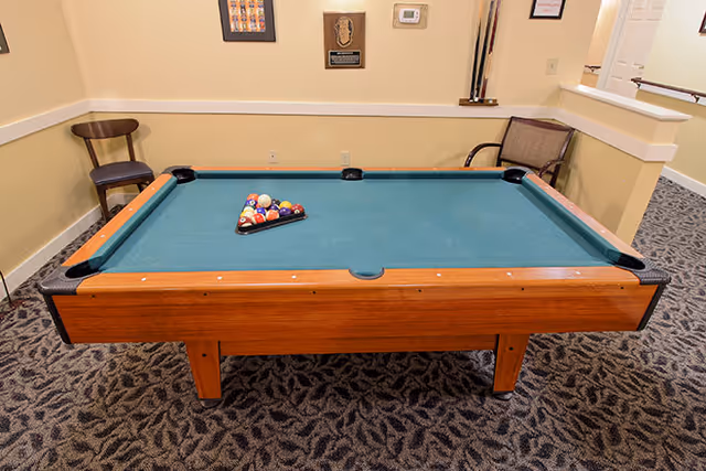 A pool table with a green felt surface and a full rack of billiard balls arranged in a triangle in the center. The table is set in a carpeted room with beige walls, a wooden chair to the left, and a cushioned chair to the right. There are plaques and a thermostat on the wall behind the table.