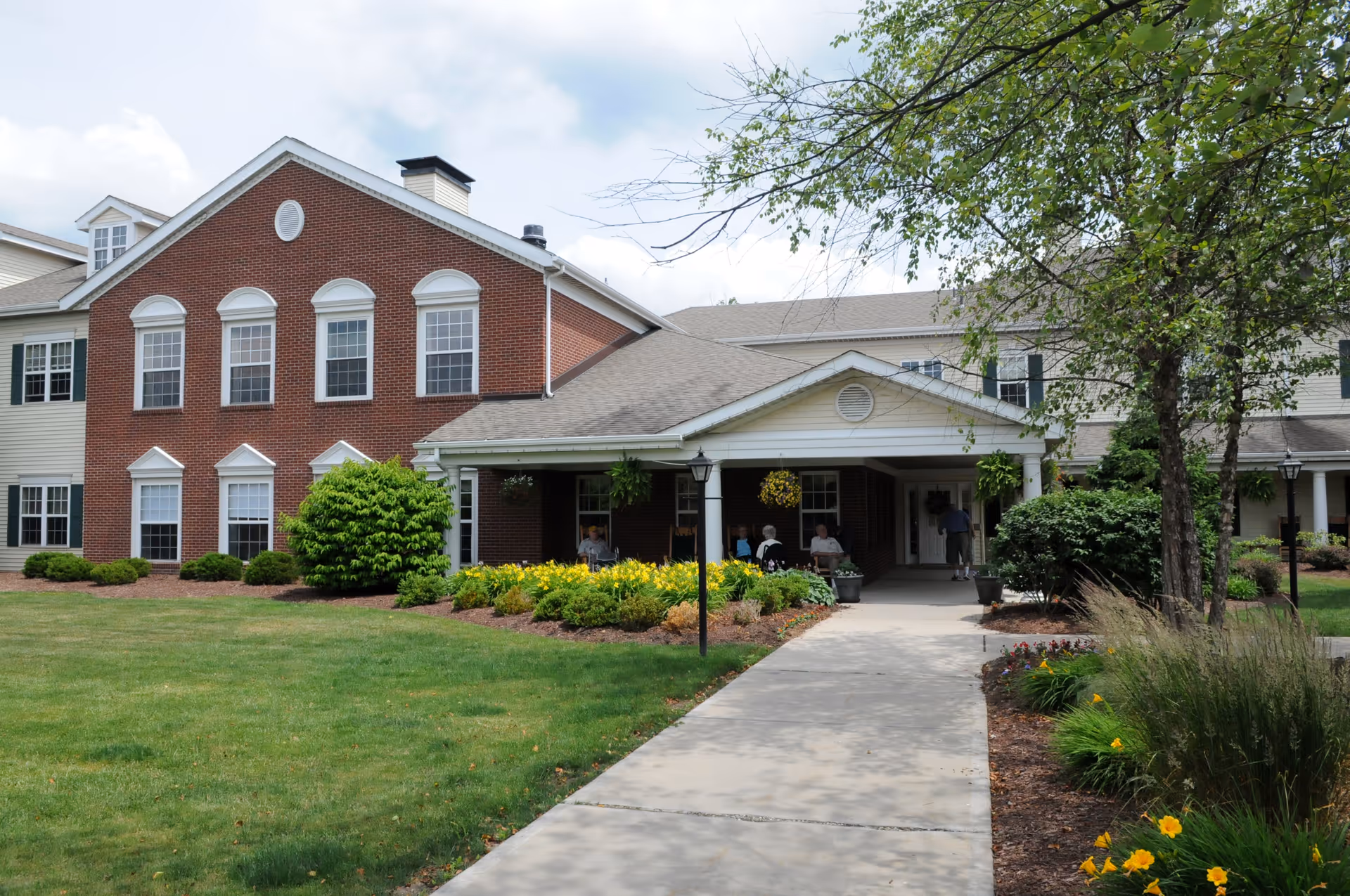 Exterior view of a senior living facility named Concordia of Cranberry, showing a brick and siding building with multiple windows, a covered entrance with people sitting and walking, surrounded by green lawn, bushes, flowers, and trees under a partly cloudy sky.
