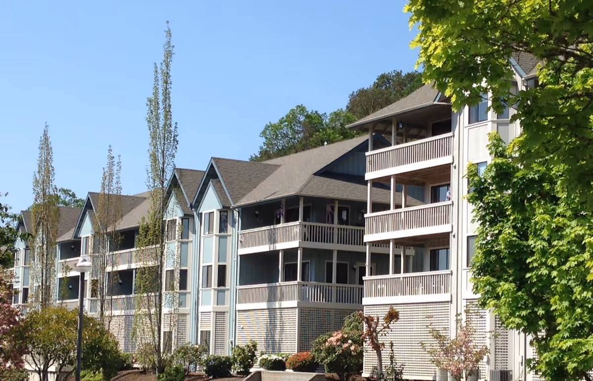 Exterior view of a multi-story residential building with balconies, surrounded by trees and landscaping under a clear blue sky.