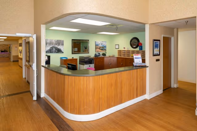 Reception area inside Granite Creek Health and Rehabilitation featuring a wooden curved front desk with office equipment and paperwork behind it, light green and beige walls, framed pictures, and a hallway leading to other rooms.