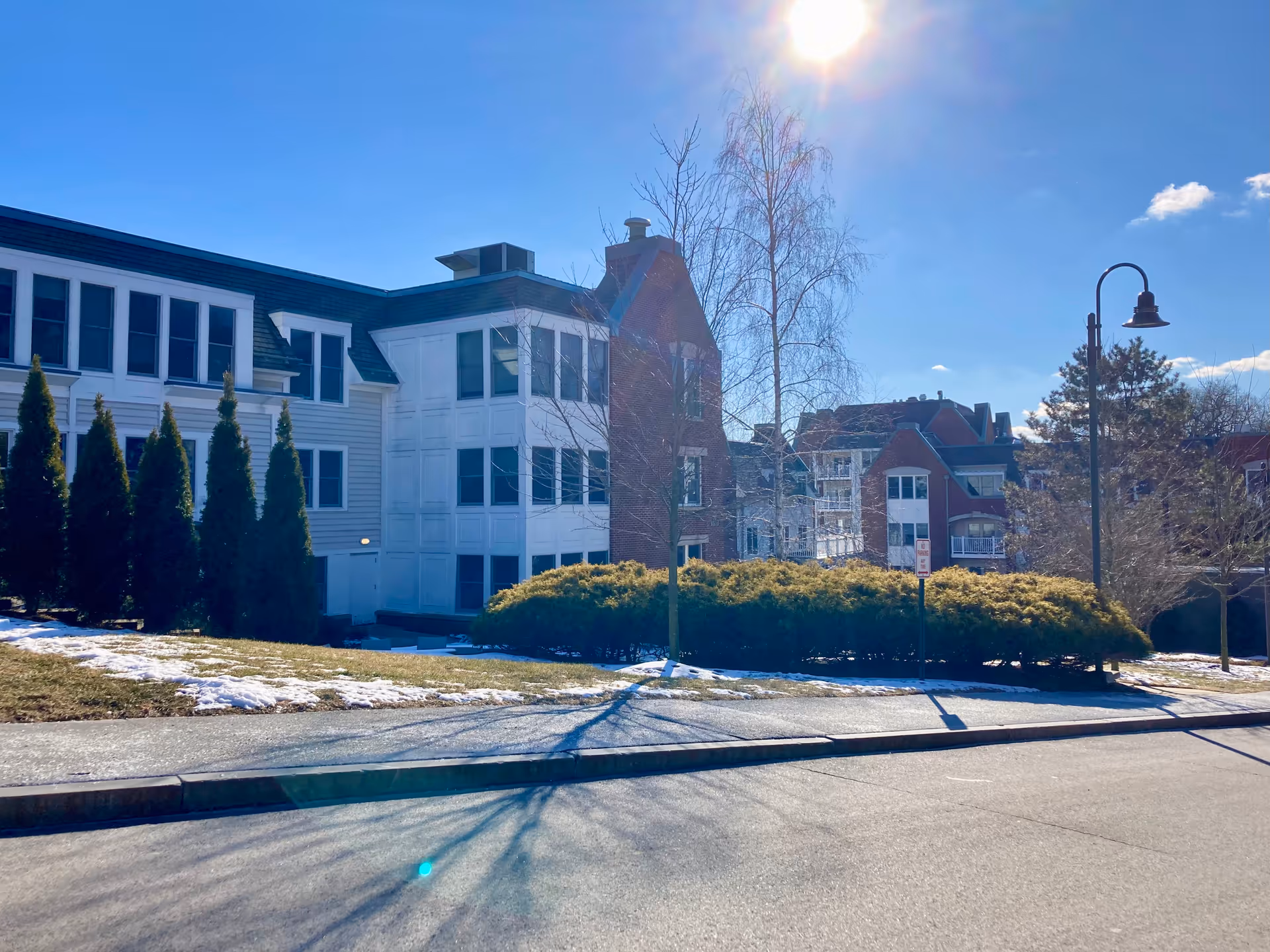 Exterior view of a senior living facility building with white and red brick walls, multiple windows, and a sloped roof under a clear blue sky with the sun shining brightly. There are some evergreen trees and bushes in front of the building, patches of snow on the grass, and a street lamp on the sidewalk.
