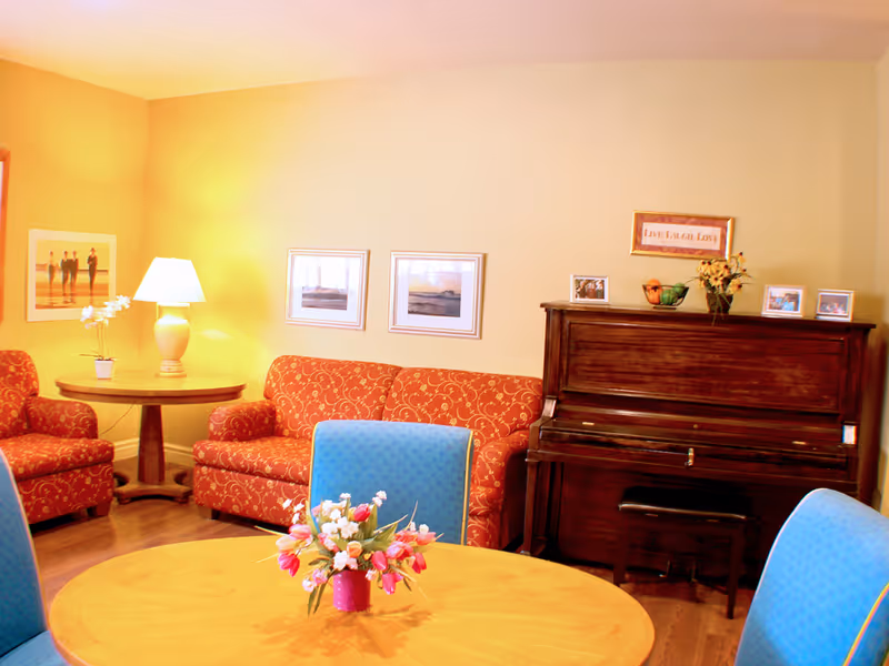 A cozy living room with a red patterned sofa and matching armchair, a wooden piano with framed photos and a 'Live Laugh Love' sign on top, a round wooden table with a flower arrangement, and blue cushioned chairs. The walls are painted light yellow and decorated with framed pictures.