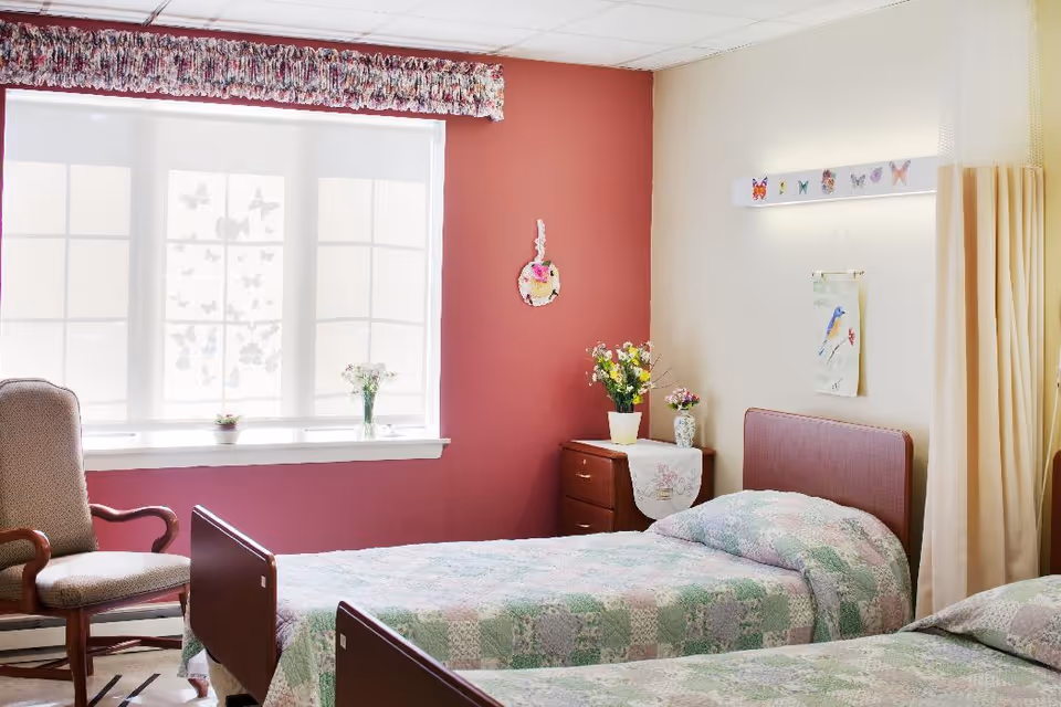 A bright and cozy nursing facility bedroom with two single beds covered in pastel patchwork quilts. There is a wooden nightstand between the beds with flower vases on top. A cushioned armchair is positioned near a large window with sheer white curtains and a floral valance. The walls are painted in a combination of soft beige and muted red, decorated with butterfly-themed artwork and a small hanging ornament.