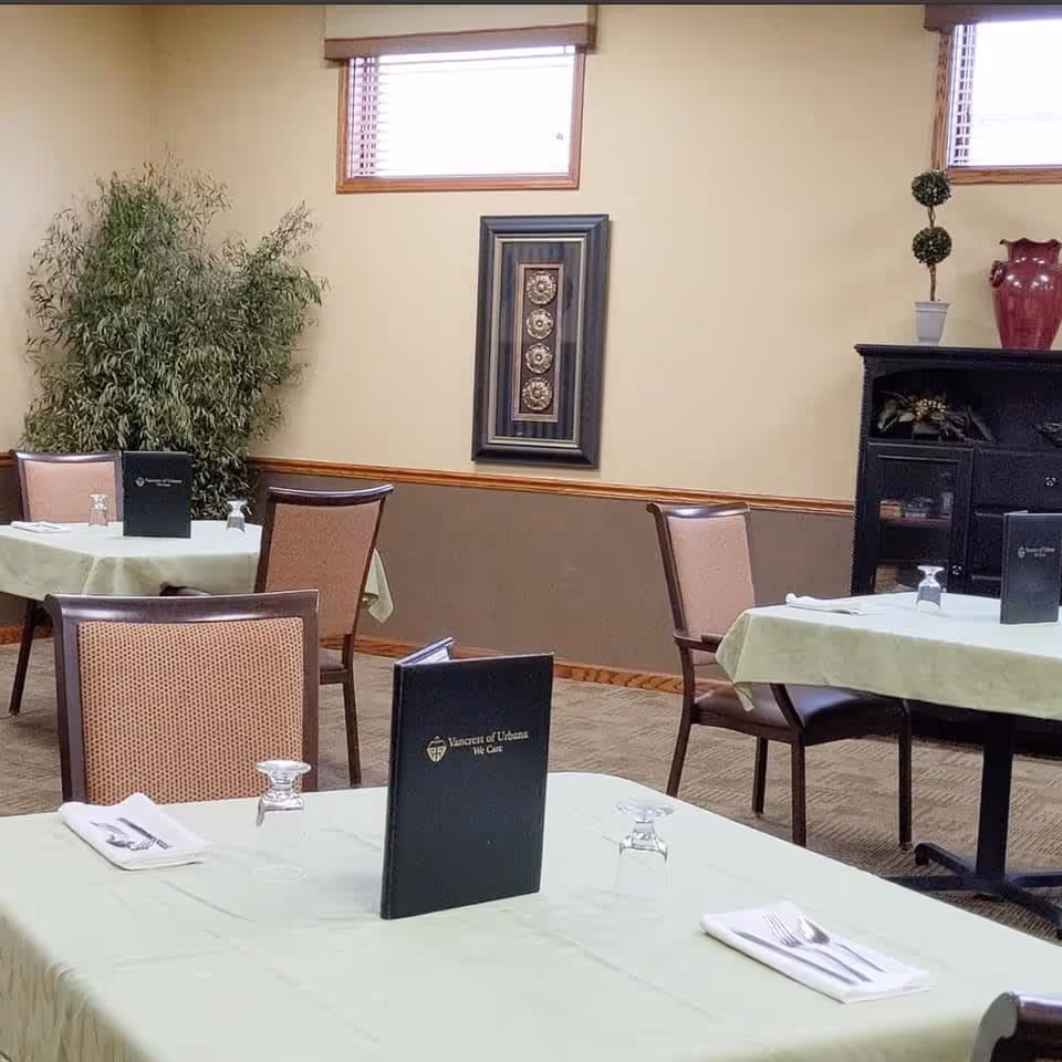 Dining room with tables covered in light green tablecloths, each set with a menu, upside-down drinking glasses, and neatly folded napkins with silverware. The room has beige walls with a brown lower half, two small windows near the ceiling, a framed decorative piece on the wall, a large potted plant in the corner, and a black cabinet with decorative items on top.