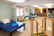 A physical therapy or rehabilitation room with a blue therapy table, exercise balls, cones, parallel bars for walking practice, and various exercise equipment. The room has a window with curtains, light-colored walls, and a polished floor.