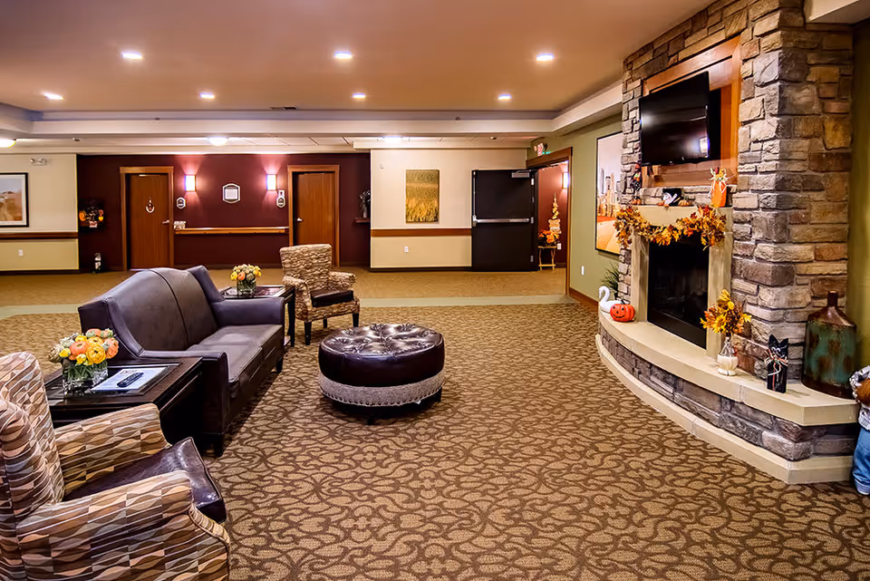 A cozy senior living facility common area with patterned carpet, a stone fireplace decorated with autumn-themed ornaments, a mounted flat-screen TV above the fireplace, a gray sofa, two patterned armchairs, a round leather ottoman, and side tables with flower arrangements. The walls are painted in warm tones with wooden doors and framed artwork.