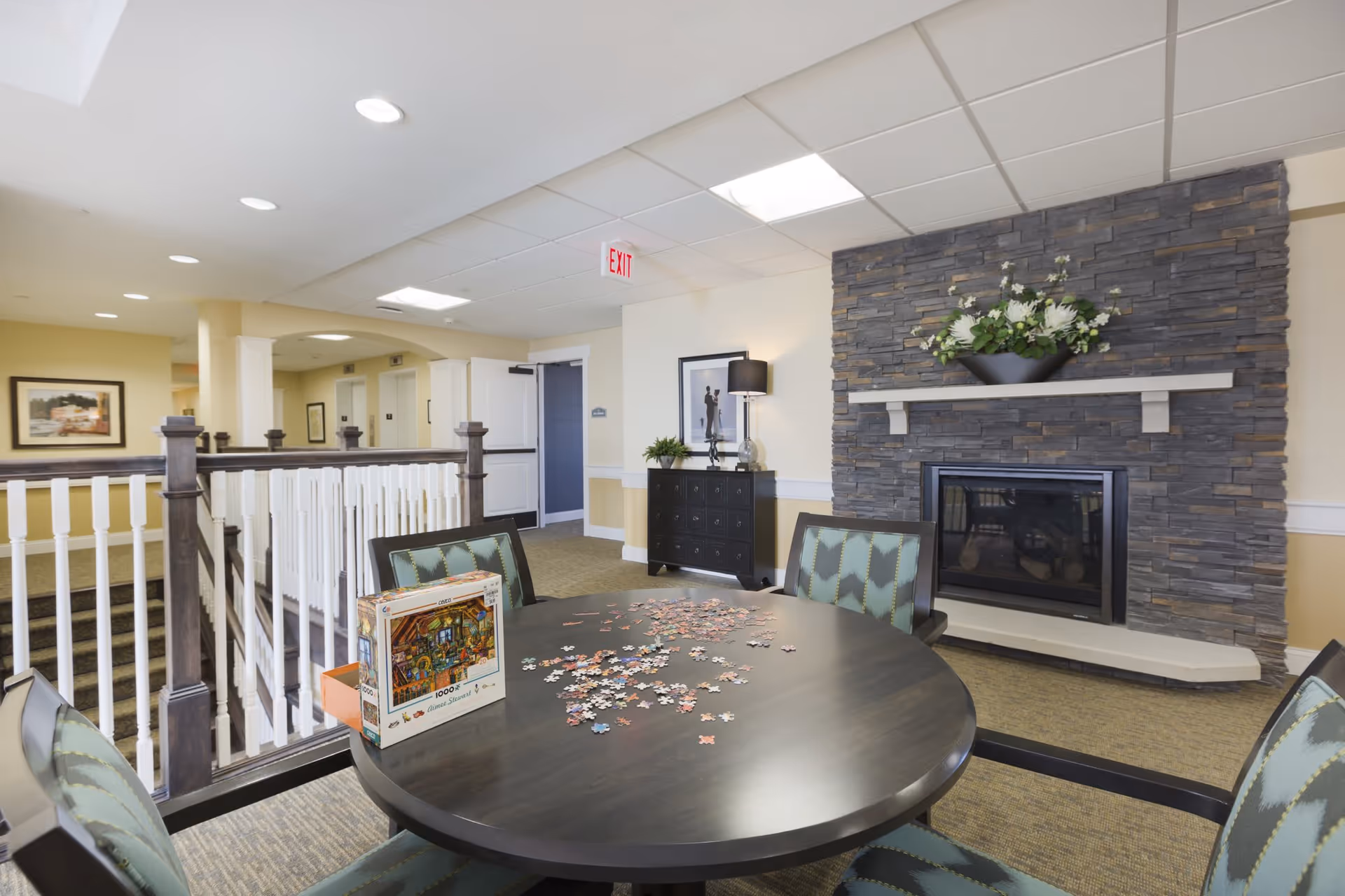 A cozy common area featuring a round table with a partially completed jigsaw puzzle and puzzle box on it, surrounded by four cushioned chairs. Behind the table is a stone fireplace with a white mantel decorated with a flower arrangement. The room has beige walls, carpeted floor, and a staircase with white and dark wood railing leading to another level. There is a black dresser with a lamp and framed artwork on the wall near the fireplace.